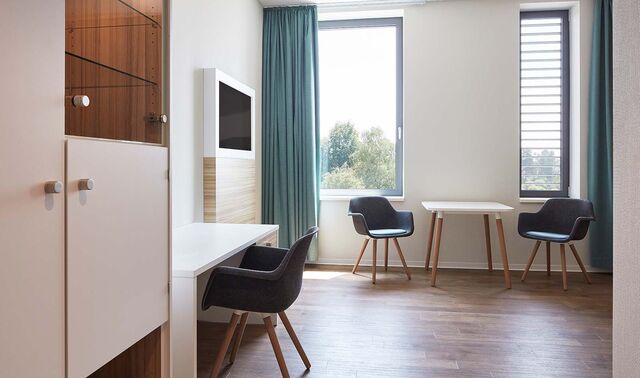 Ein Patientenzimmer in dem sich einige Stühle sowie einen Schreibtisch, einen Schrank und einen Fernseher befinden.